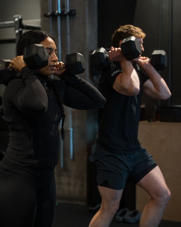 A client lifting dumbbells next to her SHĀB coach